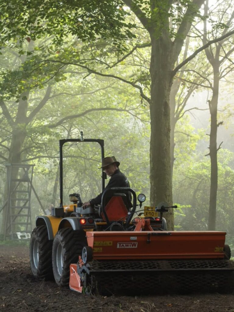Man van Wildbeheer WBE Broek en Duin op een oranje tractor die met een grondbewerker de bodem losmaakt in een bosrijke omgeving met zonlicht tussen de bomen.